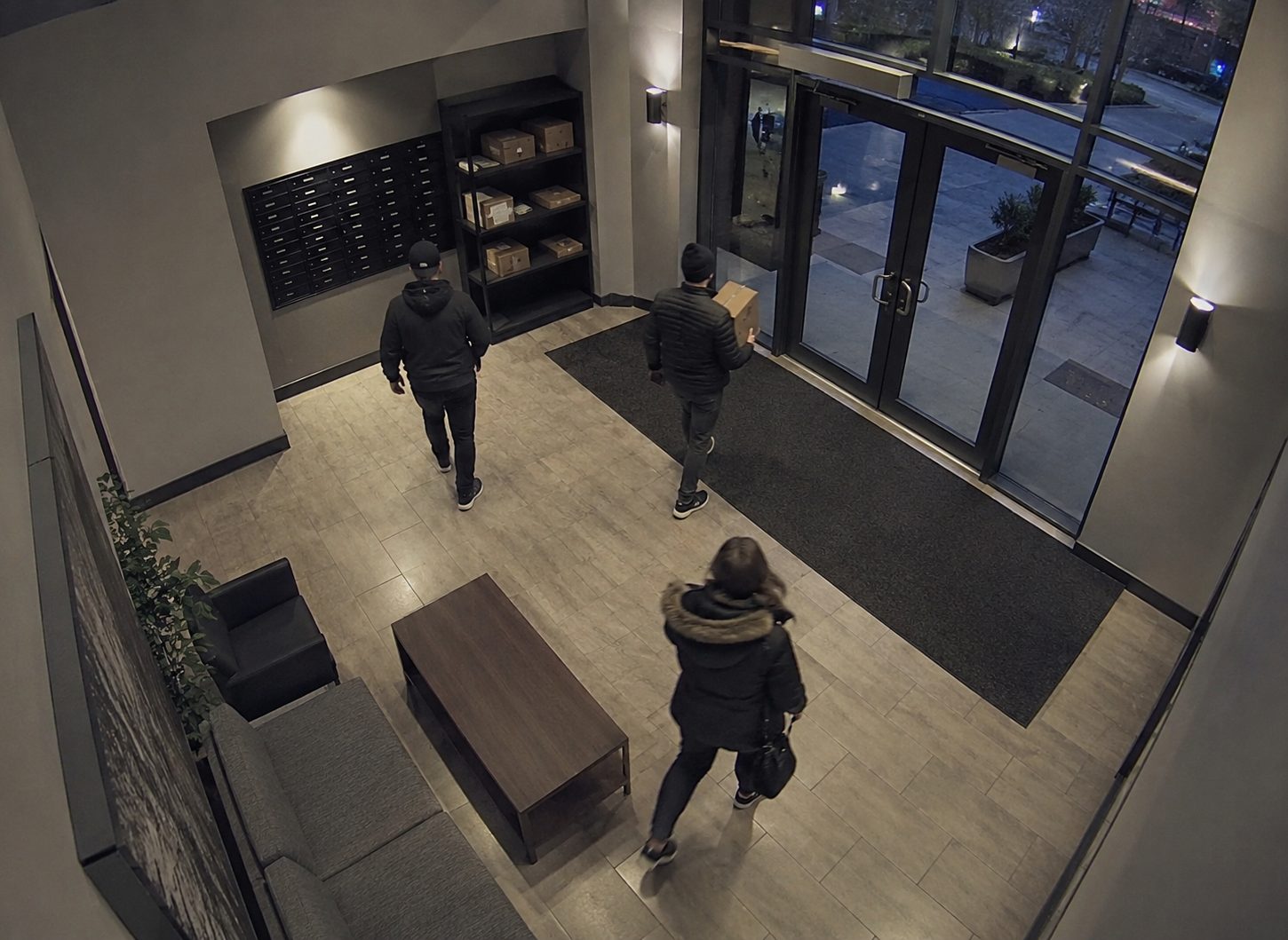 A security-camera view of an apartment lobby at night, residents crossing the floor and a parcel carried past the mailboxes and package shelves.