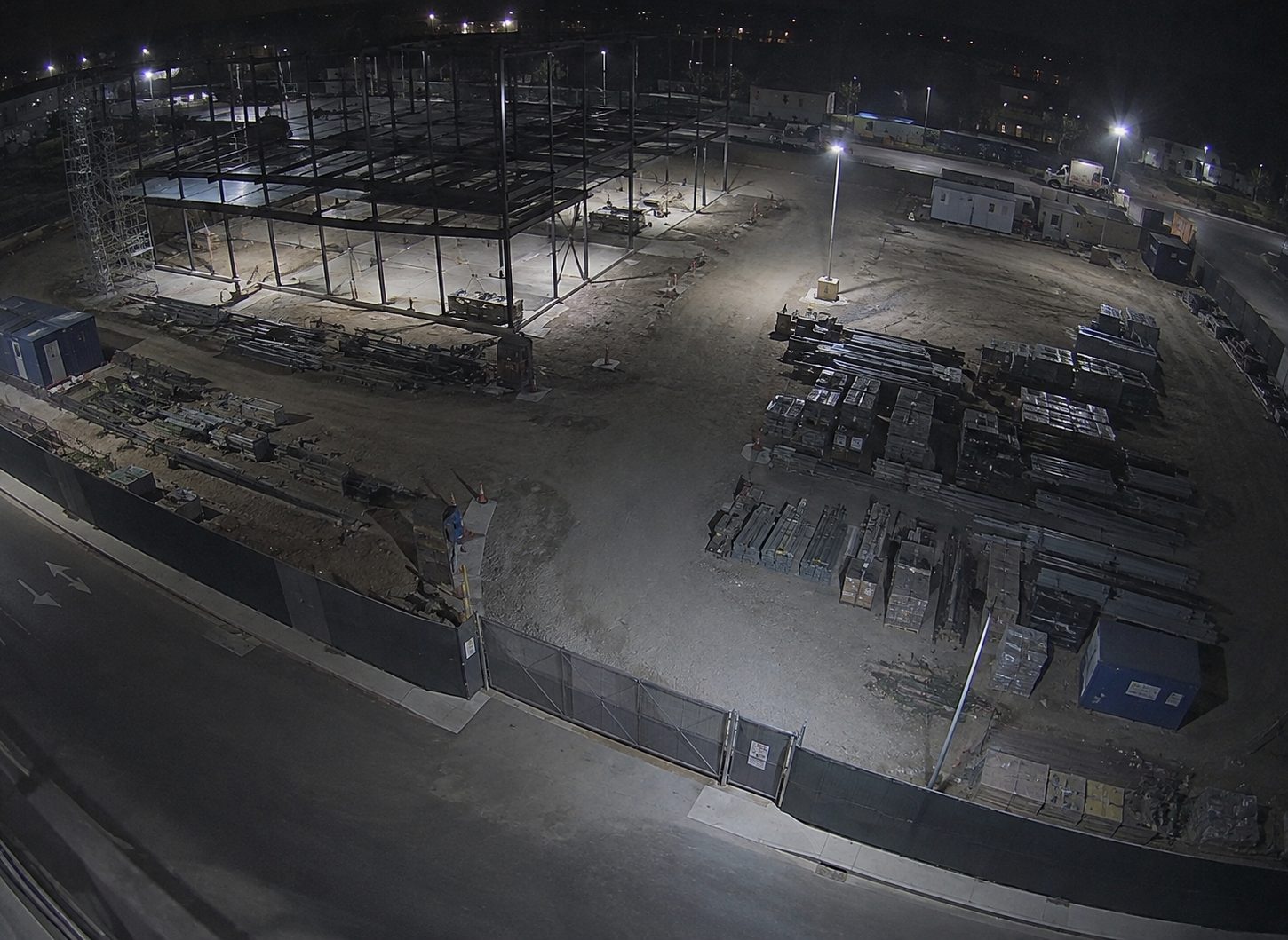 A night view of a construction site: a half-built steel frame, stacked materials and a site cabin behind perimeter fencing.