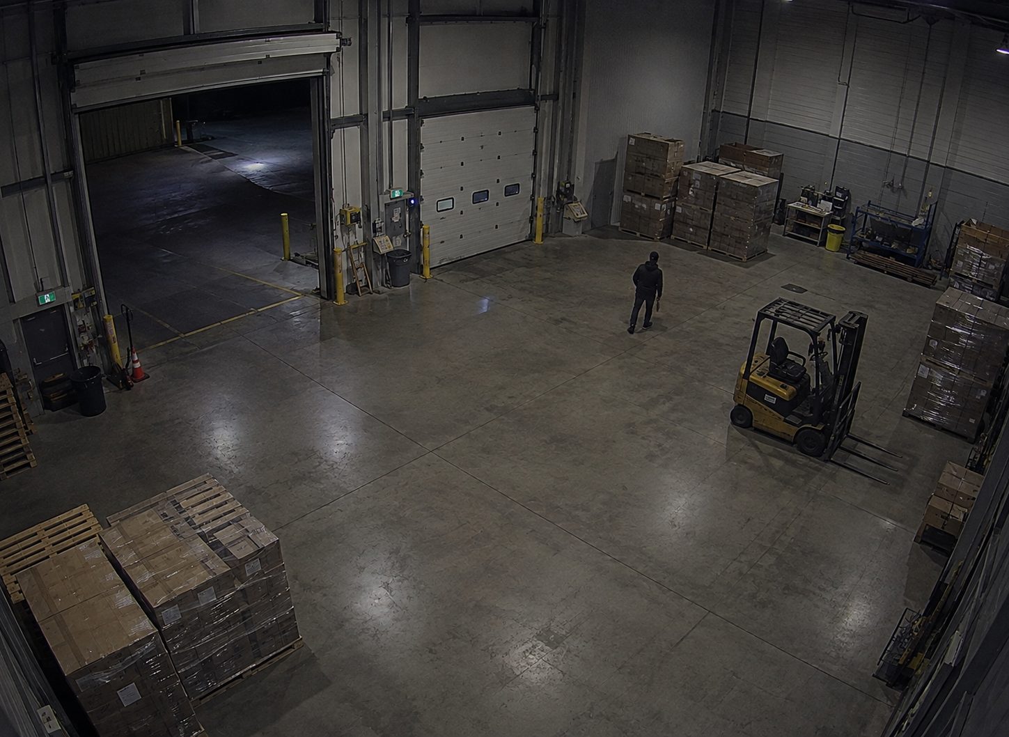 A dim warehouse at night, a lone figure standing on the floor beside a forklift among stacked pallets.