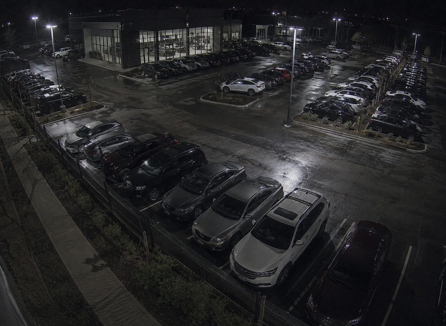 A car dealership lot at night, rows of vehicles parked under floodlights outside a lit showroom.