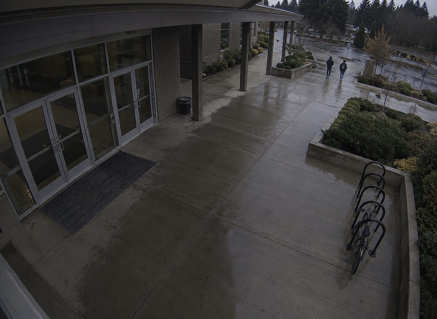 A covered walkway and bike rack at an institutional building entrance in the wet, two people walking toward the car park.